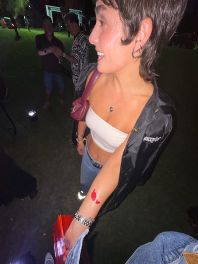 The photograph, taken in an outdoor setting, depicts a young woman in the foreground. She is wearing a white tube top, blue jeans, and a black jacket, with a burgundy purse slung across her chest. She has a silver necklace, earrings, and a watch on her wrist, along with what appears to be a red temporary tattoo on her arm. The setting appears to be at night, possibly in a park or grassy area, with a group of people in the background. One man in the background is wearing a patterned shirt. There is a lit-up object that appears to be a box, possibly containing food or other items, being held by the woman. The location could be Coachella, United States of America, though there is no specific indication in the image itself. The environment appears to be low-lit, suggesting an outdoor event or gathering.