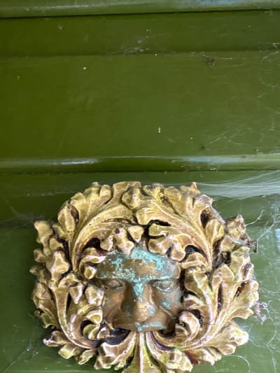 A close-up, vertical shot depicts an aged, ornate metallic decoration mounted on a dark olive-green painted surface, likely a door, in Stiperstones, United Kingdom. The decoration is a relief sculpture of a human face, characteristic of a "Green Man" motif, with foliage surrounding and intertwining with the facial features. The metallic element, appearing to be brass or bronze, exhibits significant verdigris (green patina) on the face and some of the leaf details, alongside areas of original golden-bronze color and darker tarnish. The painted surface behind the ornament shows horizontal panel lines and is covered with extensive, fine cobwebs and scattered dust particles, indicating outdoor exposure and lack of recent maintenance. No people, subjects, actions, or interactions are visible.