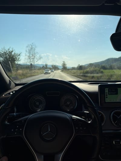 The image is a first-person view from the driver's seat of a Mercedes-Benz car on a sunny day. The steering wheel, dashboard with a navigation screen, and part of the car's interior are visible. The car is driving on a paved road that winds through a rural landscape with trees and hills. Two cars are visible ahead on the road. The navigation screen displays a map and route information, indicating a destination of Sofia, Bulgaria. The time is displayed as 10:44 and the distance to the destination is 12 km. The location is identified as Vasilkovska mahala, Bulgaria, consistent with the rural setting and the displayed route to Sofia. The sun is shining brightly, creating glare on the windshield.