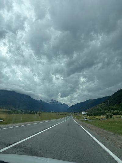This is a photograph taken from inside a vehicle, looking out at a mountain landscape. The image is oriented vertically, with the road and the right side of the landscape appearing on the right of the frame, and the sky and the left side of the mountains on the left.
The subjects include a paved road with a white line down the center, disappearing into the distance. To the left of the road is a grassy field, with some scattered trees and buildings. Further to the left, a large, dark mountain range rises steeply, its upper slopes partially obscured by dramatic, heavy clouds. The clouds are a dominant feature, filling the left side of the image with various shades of gray, indicating overcast or stormy weather. There are glimpses of snow-capped peaks on the far left, suggesting a high-altitude location.
The scene appears to be a scenic drive through a mountainous region. The presence of the road and the distant buildings suggests a populated area, likely a village or settlement in a valley. The time of day is difficult to determine precisely due to the cloud cover, but the lighting is diffused, typical of an overcast day.
Notable details include the texture of the clouds, the dark, imposing presence of the mountains, and the well-maintained road. There are a few indistinct structures visible in the distance along the roadside, and some utility poles are also present. A white vehicle is partially visible in the lower right corner, indicating the perspective is from a car. The overall mood is somewhat dramatic and imposing, due to the heavy clouds and the scale of the mountains.
There is some text visible on a sign in the middle ground on the left side of the road, appearing as a blue and white advertisement. The location context is Верхняя Теберда, Russia, which is a mountainous region known for its natural beauty and alpine climate.
