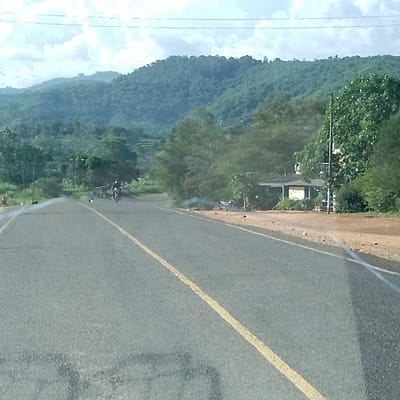 The image displays a two-lane asphalt road with a solid yellow center line, situated within the Kola ward, Kenya. A single person operates a motorcycle, traveling away from the camera in the left lane. To the right of the road, a dirt shoulder precedes a dense area of green trees and foliage, partially obscuring a single-story building with a light-colored roof and a utility pole. The background features verdant, tree-covered hills or mountains under a partly cloudy sky, with power lines visible overhead. The overall environment appears rural or semi-rural.