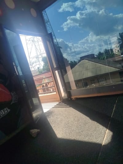 The image captures a view from inside a building in Uselu, Nigeria, looking outwards through a large window and an open doorway. The scene is characterized by strong sunlight and shadows.
The interior foreground is dominated by dark surfaces, likely furniture or walls, with a noticeable red object partially visible on the left, possibly a cushion or part of a chair. A small, grey rock rests on the dark carpeted floor in the lower-left quadrant of the image. Sunlight streams in from the right, casting a distinct triangular shadow across the floor.
Through the open doorway and the window, the exterior environment is visible. The sky is a bright blue with scattered white clouds. In the distance, a large metal structure, likely a power transmission tower, is prominent against the sky. Below this structure, there are buildings with corrugated iron roofs and what appear to be arched windows on one of them. Palm trees and other foliage are also visible, indicating a tropical or subtropical climate. The overall impression is one of a bright, sunny day with a mix of urban and natural elements visible through the open architectural spaces.
