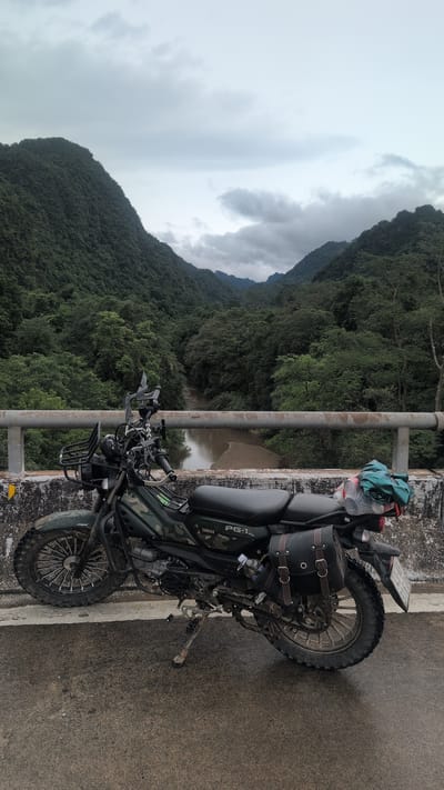 A motorcycle is parked on a bridge overlooking a river valley. The motorcycle is dark green with camouflage accents and has saddlebags and a basket on the front. It is facing to the right. The bridge has a concrete barrier and a metal railing. The valley is covered in lush green trees, and the sky is overcast. The location is Thượng Trạch Commune, Vietnam.