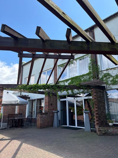 An exterior shot shows a covered seating area in front of a building with large windows. A wooden pergola with thick beams extends over the paved patio area. The building has a light-colored facade and is partially covered in green ivy. To the left, a table with chairs is set up under a large white umbrella. In the center, a set of glass doors leads into the building, with two tall grey planters flanking the entrance. The ground is paved with brickwork, and shadows from the pergola create a patterned effect. In the background, a clear blue sky with scattered clouds is visible. The building appears to be part of a hotel or restaurant, possibly located in the city of Duszniki, Poland.