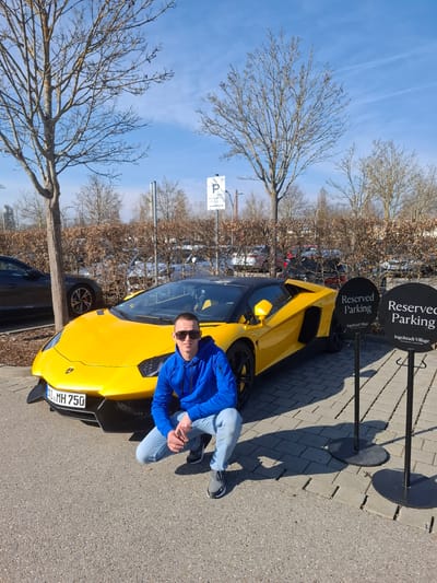 A male individual is observed squatting directly in front of a bright yellow Lamborghini Aventador SVJ in a parking area at Ingolstadt Village, Ingolstadt, Germany. The man, wearing a royal blue hooded sweatshirt, light blue jeans, and dark athletic shoes, has dark sunglasses obscuring his eyes and is looking straight towards the camera. The Lamborghini features a black roof, black multi-spoke wheels, and a German license plate displaying "IN.MH 750" on its front bumper.
To the right of the vehicle, two black circular signs mounted on poles read "Reserved Parking" with "Ingolstadt Village The Bicester Collection" printed below. The immediate ground surface where the man is positioned and the car is parked consists of light grey interlocking pavers, transitioning to asphalt in the closer foreground. In the background, several deciduous trees stand bare, indicative of winter or early spring, alongside a leafless brown hedge. A dark grey sedan is partially visible parked behind and to the left of the Lamborghini. The sky is clear and blue, showing faint contrails, suggesting a bright, sunny day.