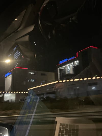 The media file captures a rotated (90 degrees clockwise) nighttime scene viewed from inside a vehicle, dominated by reflections on the glass. The dark environment is illuminated by various artificial lights.
Inside the vehicle, a portion of a steering wheel is visible in the bottom-left corner. Faint reflections of a person's hands and upper body are discernible, particularly on the right side near a reflected laptop keyboard and screen. A reflected display, likely a smartphone or car screen, shows text in the bottom-center.
Through the glass, several buildings are visible in the background, featuring illuminated windows with white and blue lights. A prominent vertical line of evenly spaced orange-yellow lights stretches across the right side of the image. Additionally, red linear lights appear at the top and bottom edges, possibly outlining architectural features or signage.
The activity depicts a static moment, likely from a parked or stopped vehicle at night. The visible text on the reflected screen, partially upside down, includes "S-mobile", "1. Σύνδεση" (1. Login), "2. Εγγραφή" (2. Register), and "3. Ξεχάσατε τον κωδικό;" (3. Forgot password?). These Greek phrases, combined with the overall setting, place the scene in Agios Stefanos, Greece.