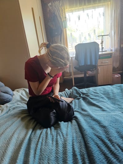 A young woman with blonde dreadlocks, wearing a maroon t-shirt and black pants, sits on a blue-gray textured comforter on a bed in what appears to be a bedroom. She is hunched over, with her head bowed and her right hand covering her face. She holds a black electronic device in her left hand. To the left of the bed are two large, gray, cushion-like objects. Behind the bed, a wooden wardrobe with gold handles is visible on the left, and on the right, a window with sheer curtains reveals trees and daylight. A desk with a chair and a pink wastebasket are also present near the window. The scene is indoors and well-lit by natural light.