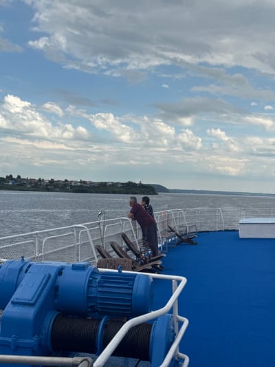 The image depicts a view from the deck of a boat on the Volga River near Ikskoe Ust'e, Russia. In the foreground, a large blue winch mechanism is visible, with black rope wound around its spool. The deck itself is covered in vibrant blue carpeting.
In the mid-ground, a white railing separates the deck from the river. Beyond the railing, two individuals are standing and looking out at the water and the opposite shore. One person, wearing a maroon shirt, is leaning on the railing, while the other, in a dark patterned shirt and dark pants, stands beside them. Several wooden deck chairs are also visible, some folded and some open.
The far bank of the river is lined with a strip of trees and a settlement of houses with colorful roofs, characteristic of a Russian village. The water of the Volga River is choppy, reflecting the cloudy sky. The sky itself is a mix of blue and grey clouds, with some brighter white clouds scattered throughout, suggesting a partly cloudy day. The overall impression is one of a leisurely moment on a river cruise, with the natural landscape and a glimpse of rural life.