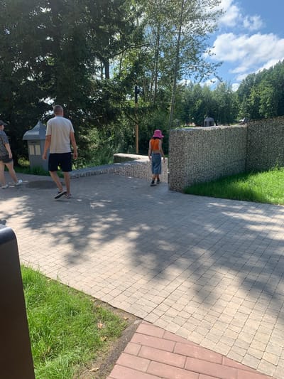 The image shows a paved walkway in a park-like setting. On the left, a man in a light-colored t-shirt and dark shorts walks away from the camera. Beside him, another person, partially visible, is also walking away. Further down the path, a young person wearing an orange crop top, denim shorts, and a pink bucket hat is walking away from the camera. The walkway is made of light-colored interlocking pavers, with a section of red brick pavers in the foreground. To the right of the walkway is a gabion wall filled with stones, with green grass beyond it. In the background, there are trees and a blue sky with white clouds. A dark trash receptacle is visible in the lower left corner. There is no information in the image to indicate the location is Arbuzov-Baran, Russia.