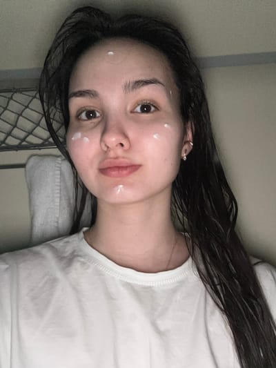 A young adult female is depicted in a close-up, front-facing shot inside a train car, identified as being in Kabinetnoye, Russia. She has dark, shoulder-length hair that appears damp and a neutral facial expression, looking directly at the camera. She wears a plain white t-shirt, a thin gold chain necklace, and a small gold hoop earring in her right ear. Her face features several distinct, unblended white dots of a creamy substance on her forehead, both cheeks, and chin. To her left, in the background, a metal mesh structure is visible, consistent with a bunk or luggage rack. Below this, a white towel hangs draped over a horizontal metal bar. The background to her right and above consists of a plain, light-colored interior wall or ceiling, featuring a faint horizontal seam or stripe. The lighting is internal and soft.