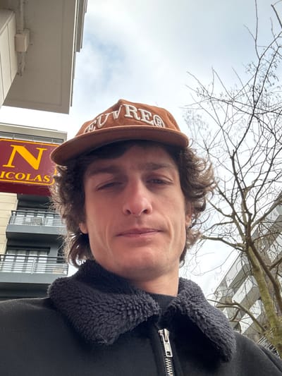 A male individual is captured in an upward-angled, close-up photograph, standing outdoors on an overcast day. He is positioned in front of a commercial and residential streetscape at 10 Rue de Paris, Joinville-le-Pont, France. The subject has medium-length, wavy brown hair and is wearing a brown corduroy baseball cap with the word "OUVREA" embroidered in white on the front. He is also dressed in a black jacket featuring a prominent black faux fur or fleece collar and a visible silver-toned zipper down the front. His expression is neutral as he looks directly at the camera.
In the background, to the subject's left, a segment of a red commercial sign displaying "N NICOLAS" in large yellow capital letters is clearly visible, identifying a Nicolas wine shop. Behind and below this sign, a multi-story modern building with dark-framed windows and dark-railed balconies is partially visible. To the subject's right, bare branches of a deciduous tree extend upwards against a bright, cloudy sky, indicating a winter or early spring season. Another building with a reflective facade is faintly discernible behind the tree branches.