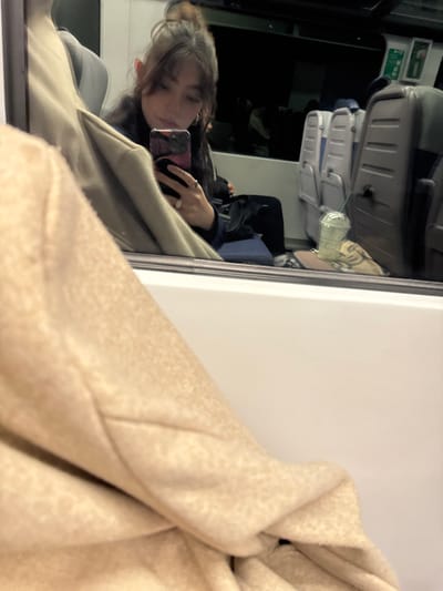 A person is reflected in a window while holding a smartphone, seemingly taking a photo of themselves. The reflection reveals they are seated on what appears to be a bus or train. Rows of seats are visible behind them. The right side of the image is dominated by a large, beige hooded jacket, obscuring some of the background. The jacket's fabric has a textured appearance.
