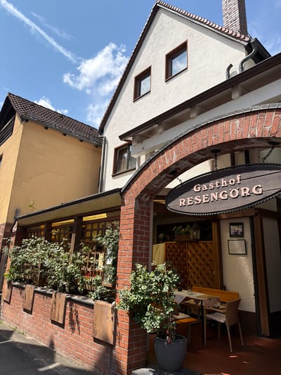 The image shows the exterior of a building, identified as "Gasthof RESENGÖRG" in Ebermannstadt, Germany. The building has a light-colored facade with a tiled roof and a prominent brick archway. Hanging from the archway is a dark wooden sign with the name of the establishment.
To the left of the main building, a yellow-colored building with a tiled roof is visible. The area beneath the archway appears to be an entrance to the establishment, possibly a restaurant or inn, with an outdoor seating area. This seating area includes wooden tables and chairs, covered by a white tablecloth, and surrounded by potted plants and greenery. The ground is paved with reddish tiles.
The scene is set on a sunny day, as indicated by the bright lighting and shadows cast on the ground. The sky is blue with scattered white clouds. The architecture suggests a traditional European style.