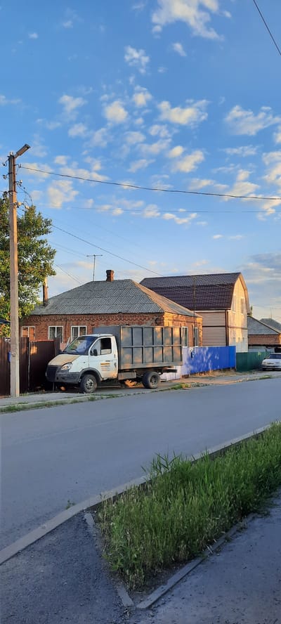 The image depicts a residential street scene in Shakhty City District, Russia, during daylight with a bright blue sky dotted with white clouds. In the foreground, a paved road runs diagonally from the bottom left to the upper right. A strip of overgrown green grass runs along the right side of the road, separated by a concrete curb.
To the left of the road, a light-colored truck with a dark metal cargo container is parked on the side. The truck appears slightly weathered. Behind the truck, a brick house with a grey, corrugated roof is visible. A brown wooden fence stands in front of the house. A tall utility pole with electrical wires is situated to the left of the fence and house.
Further down the street, another house with a darker, pitched roof and light-colored siding is present. A bright blue fence is situated between the two houses. A white car is parked on the right side of the street, further down from the blue fence. The overall environment suggests a quiet, rural or suburban residential area. No people are visible in the image.