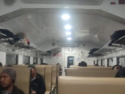 The interior of a train car in Kecamatan Beji, Indonesia at coordinates -7.581 latitude, 112.718 longitude. The car is sparsely populated with a few passengers seated on the tan-colored seats. There are overhead luggage racks with luggage in them. There are several overhead lights, some of which are on. The walls are white and the ceiling is white. There is a door at the end of the car. The car appears clean and well-maintained. The atmosphere is calm and peaceful.