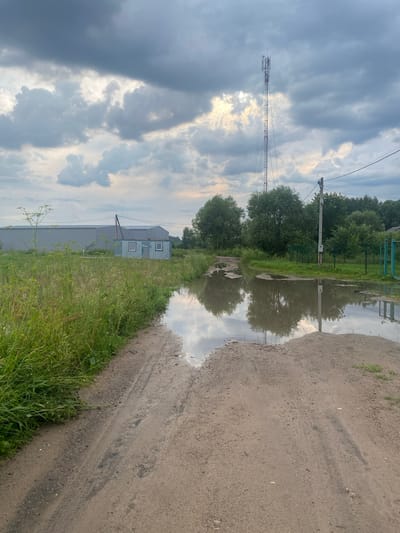 The image depicts a rural scene in the city of Zavolzhsky, Russia, characterized by a dirt road with large puddles reflecting the cloudy sky. To the left, a field of tall green grass and some scattered wild plants is visible, adjacent to a long, grey, industrial-style building with a smaller, blue, shed-like structure attached to its side. This structure has two windows. In the background, a tall telecommunications tower stands prominently against the overcast sky, with power lines and poles also present. Further to the right, a line of lush green trees forms the background, with a green metal fence running along the edge of the property. The overall impression is one of an overcast day in a semi-industrial or agricultural area.