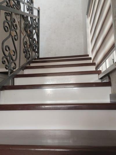 This is a low-angle, eye-level shot of a staircase, viewed from the bottom looking up. The staircase has dark brown wooden steps with white risers and is bordered by a dark metal railing with decorative scrollwork and gold accents on the left. The wall on the left is textured and light-colored, while the wall on the right features horizontal blinds in shades of white and brown. The scene appears to be indoors, likely in a residential setting, and the lighting suggests it is daytime. There are no people or any discernible text visible in the image. The overall impression is one of a well-maintained interior staircase.