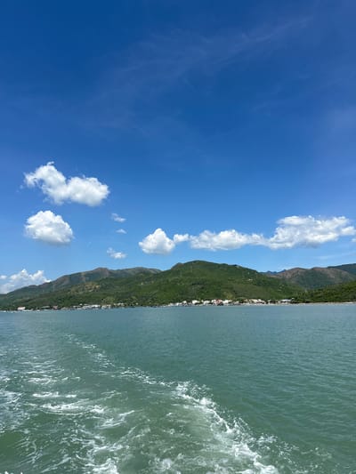 The image depicts a scenic coastal view. On the left, a vast expanse of bright blue sky is dotted with fluffy white clouds, some wispy and others more substantial. A steep, verdant mountainside runs along the coastline, densely covered in green vegetation. A small, linear settlement with white buildings is visible nestled along the base of the mountain, bordering the water. The right side of the image is dominated by the ocean, which has a greenish-blue hue and shows signs of gentle movement with small waves and white foam. The photograph appears to be taken from a vantage point on the water, looking towards the shore. The bright sky and clear visibility suggest it is daytime, likely mid-morning or afternoon, with fair weather. The scene evokes a sense of tranquility and the natural beauty of a coastal landscape. There is no visible text in the image.