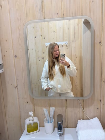 The image shows a mirror selfie taken by a woman in what appears to be a bathroom or dressing area, set in Vozhoiskii, Russia. The image itself is rotated 90 degrees counter-clockwise, showing the subjects and objects on their sides.

The primary subject is a young woman, reflected in a rectangular mirror with rounded corners. She has long, straight blonde hair and is wearing a light cream or off-white hooded sweatshirt (hoodie) and matching sweatpants. Her left arm is visible, holding a smartphone with a light-colored case up to take the picture. She is looking at the camera with a slight smile. A notable detail on her hoodie is an illuminated square symbol, possibly a power icon or a brand logo, glowing on the front.

The setting features walls made of light-colored wooden planks, creating a rustic or cabin-like atmosphere. To the right of the mirror, a white ceramic sink and a modern chrome faucet are visible. On the sink's surface, there's a white holder containing two individually packaged dental kits, and a white and yellow liquid soap dispenser. A stack of neatly folded white towels and a white electrical cord are also visible near the sink.

Visible text includes "用心为你做 Le" and "HOTEL DENTAL KIT" printed on the packaging of the dental items, indicating they are amenities. The brand "Lersanit" is also visible on the sink or a nearby fixture. The overall impression is a well-lit, clean space.