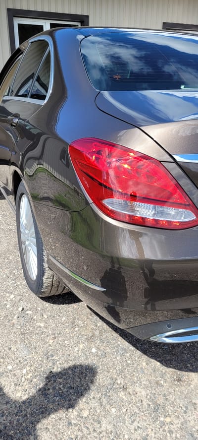 The image shows the rear passenger side of a dark brown sedan parked on a gravel surface. The car's taillight assembly is prominently featured, with its red and clear lenses reflecting the bright sky and clouds. The car's metallic paint is highly reflective, showing distorted images of the surroundings, including what appears to be a building with vertical siding and a window. The ground is covered in loose gravel and small stones, with patches of green grass visible along the edge of the car. A shadow of a person is cast on the ground to the left of the vehicle. The scene is likely outdoors, given the natural lighting and shadow. No specific landmarks or identifying features of Lielceri, Latvia are visible in the image.