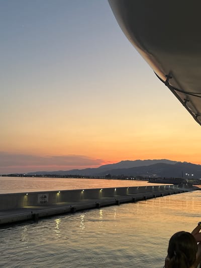 The image depicts a sunset view from a vessel in the port of Motril, Spain. The sky transitions from a pale blue at the top to vibrant orange and pink hues near the horizon, where distant mountains are silhouetted against the colorful sky. A long, illuminated breakwater extends into the water, with lights spaced along its top. The water reflects the warm colors of the sunset. In the foreground, the edge of a large, white structure, possibly part of the vessel, juts into the frame from the upper right. The back of a person's head and shoulders is visible in the lower right corner, suggesting someone is observing the scene. The overall atmosphere is serene and picturesque.