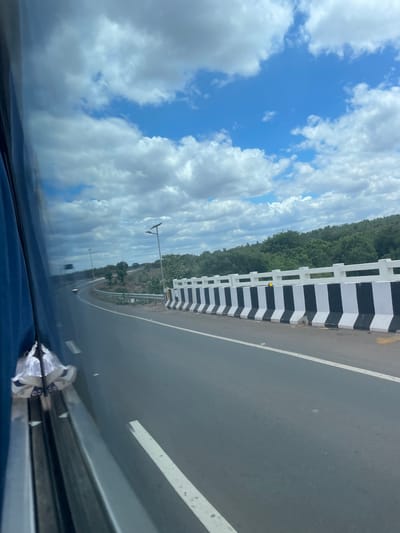 A highway runs through a landscape of hills covered in green trees under a bright blue sky with scattered white clouds. The highway has a black asphalt surface with white lane markings. A white concrete barrier with black and white diagonal stripes runs along the right side of the road. In the distance, the road curves to the left. There are streetlights visible along the side of the road. The image is taken from inside a vehicle, with a blue tinted window and part of the interior visible on the left side of the frame. There is some crumpled white paper or plastic caught on the interior of the vehicle. The location is identified as Sugali Tanda, India.