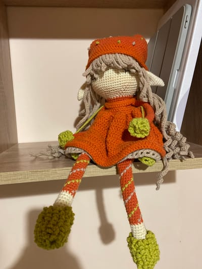 A crocheted doll with light brown hair and pointed ears sits on a wooden shelf in what appears to be a domestic environment. The doll is wearing an orange hat with small yellow and white beads, an orange dress with a ruffled hem, and striped leggings in orange, white, and yellow. It has green crocheted pom-poms on its feet and as decorative elements on its dress and hat. A silver tablet is placed behind the doll on the same shelf. The background wall is a light pinkish-beige color. There is no visible indication of the city of Aginskoye, Russia, in the image.