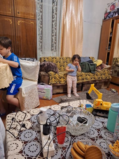 In Zeda Sazano, Georgia, an indoor scene captures a young boy, likely Caucasian, wearing a blue t-shirt and blue shorts with white text, sitting at a table covered with a yellow patterned tablecloth. To his right, a white chair is draped with a light-colored patterned fabric. In the background, a little girl, also likely Caucasian, stands on a rug in light pink pants and a light blue t-shirt, with her hand to her mouth. She is positioned in front of a sofa with a yellow and black ornate pattern. A green duffel bag and other items are on the sofa. To the right of the sofa, a toy yellow excavator is on the rug, and a small toy car is visible further to the right. A large wooden wardrobe with intricate carvings is on the far left of the image. The floor appears to be wooden. In the foreground, a table is covered with a crocheted white and grey circular patterned tablecloth. On this table, there is a white power strip with multiple outlets and electrical cords, a phone stand, a glass bowl with various small items, a tube of red cream, and a glass bowl filled with cookies. A teal water bottle and a partially visible patterned bottle are also on the table. The overall impression is a cluttered domestic environment with children and toys present.
