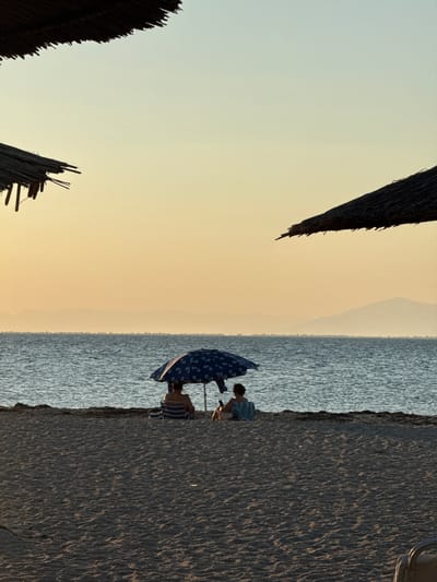In Angelochori, Greece, at dusk, two individuals sit beneath a blue patterned umbrella on a sandy beach, facing a tranquil sea. The sky is a soft gradient of pale yellow and blue, with faint mountain silhouettes visible on the horizon. Two thatched beach umbrellas frame the upper corners of the image, casting subtle shadows. The sand is textured and dappled with sunlight and shadow. One person is seated in a striped chair, while the other sits on the sand with a towel. The overall scene evokes a sense of peaceful relaxation by the sea.