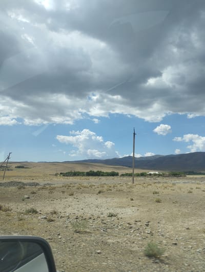 This is a landscape photograph taken from a moving vehicle in the O'xum QFY, Uzbekistan. The sky is filled with dramatic, heavy grey clouds interspersed with patches of bright blue sky and fluffy white cumulus clouds. In the distance, rolling hills and mountains are visible, appearing muted in color due to the atmospheric conditions. The foreground consists of a dry, arid landscape with sparse vegetation, rocks, and dirt. A power pole with wires runs horizontally across the scene, and another pole is visible to the left. A small cluster of trees and some low-lying structures, possibly buildings or agricultural structures, are situated on the mid-ground hills. A portion of a car's side mirror is visible in the bottom left corner of the frame, indicating the perspective of the shot. The overall impression is of a vast, somewhat desolate, and possibly remote rural environment under a dynamic sky.