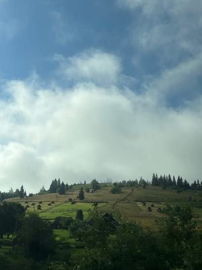 The image depicts a rural hillside landscape in Yablunytsia, Ukraine. The sky is a pale blue with dramatic, bright white clouds dominating the upper half of the frame. Below the clouds, a rolling green and golden hillside is dotted with numerous haystacks. A few scattered evergreen trees stand on the slope, and a line of taller evergreen trees forms a dense border along the crest of the hill. In the mid-ground, a wooden fence encloses green pastures. Small, dark structures, likely farmhouses or barns, are nestled within the landscape. The foreground is filled with lush, dark green foliage. The scene is a tranquil, agrarian view of the Ukrainian countryside.