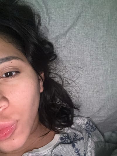 The image displays a partial view of a female individual in an indoor setting. The subject's face is positioned on the left side of the frame, angled towards the camera. Visible features include her left brown eye, the bridge of her nose, and her mouth with full, slightly parted lips. Dark, wavy hair frames her face and extends onto a light grey, textured fabric surface that occupies the right half and upper portion of the image. The fabric, consistent with bedding or a pillow, is visibly creased. The individual is wearing a light grey garment with a repeating pattern of dark snowflake-like designs, and a white layer is visible beneath its neckline. A dark olive-green fabric is partially visible in the lower right corner. The individual is lying down, looking directly into the camera. This scene is located in Atizapán de Zaragoza, Mexico.