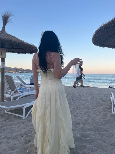 A medium shot shows a woman from behind, standing on a sandy beach in Platja D'alcúdia, Spain. She has long, dark hair adorned with a white flower and is wearing a light yellow, floor-length dress with ruffled layers. Her right hand is raised, holding a sheer white veil or fabric. In the background, two people walk along the beach near the water's edge. The sea extends to the horizon, with a coastline and hills visible in the distance. Two thatched umbrellas and empty lounge chairs are present on the sand. The sky is a clear, pale blue, suggesting it is late afternoon or early evening.