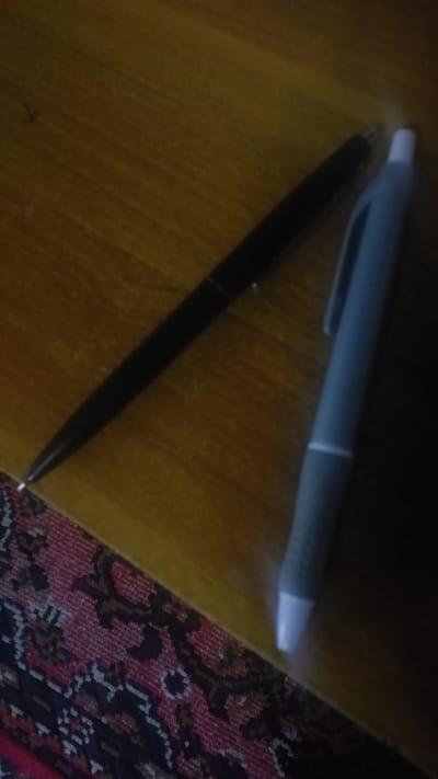 Two pens are on a wooden surface, possibly a desk or table, which is resting on a patterned rug. One pen is black and is positioned diagonally across the wooden surface, with its tip pointing towards the bottom left. The other pen is light blue or grey and is positioned to the right of the black pen, also diagonally, with its tip pointing towards the bottom right. The wooden surface has visible grain. The lighting in the image is dim, creating shadows and making some details less clear. There are no people visible in the image. The location is not specified as Ghafurov, Tajikistan.