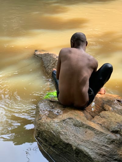 A young man sits on a large rock in a river near the town of Egbeda, Nigeria. He is facing away from the camera, with only his back visible. He wears black pants and no shirt. The water of the river appears muddy and brown in color. The rock is large and brown, with some green vegetation around it. There is a slight current in the river, which causes the water to flow around the rock and create a whirlpool effect. The man's feet are dangling in the water. The man appears to be enjoying the river. The river is located near the coordinates 7.119, 5.362.