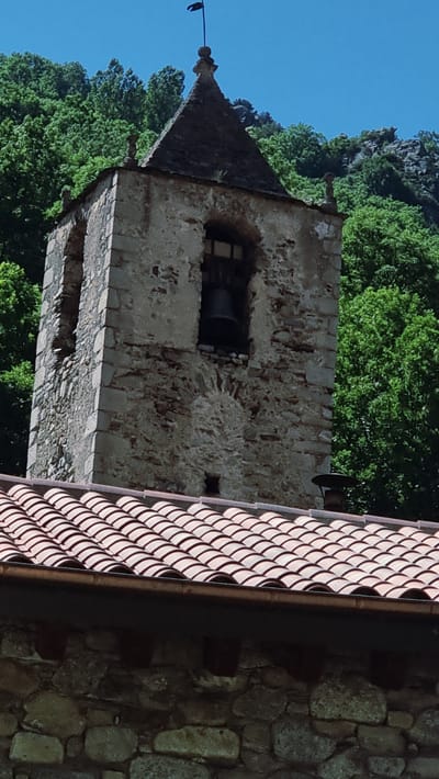 The image shows the upper portion of an old stone bell tower, with a conical dark roof, set against a backdrop of lush green trees and a clear blue sky. To the right of the tower, a section of a terracotta-tiled roof is visible, with a dark gutter running along its edge. The stone construction of the tower appears weathered and aged, with visible mortar lines and irregular stonework. A dark bell is visible through an arched opening in the tower. A thin wire or cable extends from the peak of the tower's roof, with a dark object, possibly a bird, perched on it. The scene suggests a rural or historical setting, possibly a church or castle in Setcases, Spain. The bright sky and green foliage indicate a daytime scene, likely during pleasant weather. No text is visible in the image.