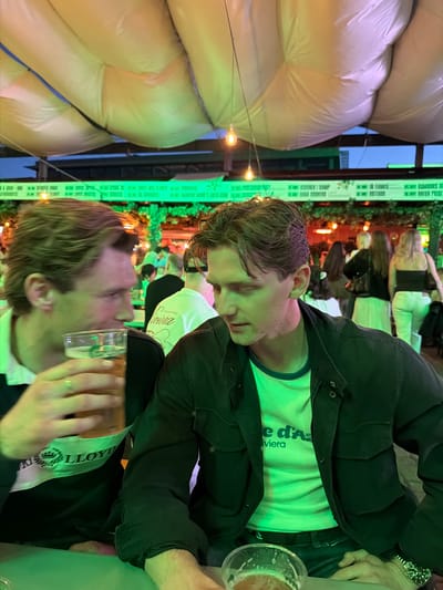 The image shows two young adult men sitting at a table outdoors. The man on the left is holding a plastic cup filled with a light-colored beverage, possibly beer, and is looking away from the camera. He is wearing a dark-colored polo shirt with a white collar and a logo that reads "LLOYD" on the chest. The man on the right is looking down and to his left. He is wearing a dark jacket over a light green t-shirt with text that appears to read "e d'A Riviera". Both men have light skin and brown hair.

The environment appears to be a lively outdoor venue, possibly a bar or restaurant. In the background, several other people are visible, and there are string lights and what look like large inflatable structures overhead. A green neon sign with text is visible above the heads of the people in the background. The overall lighting suggests it is evening. The presence of the text "Tønsberg, Norway" in the prompt indicates this scene is located in Tønsberg, Norway.