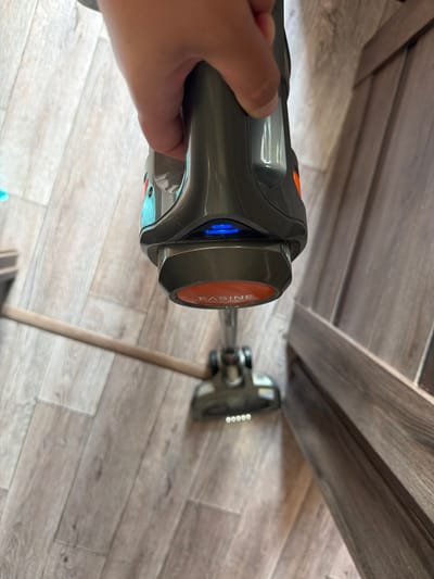 A hand is holding a grey and orange handheld vacuum cleaner with blue indicator lights and the word "EASINE" on an orange disc at the bottom. The vacuum cleaner is positioned over a light-colored wooden floor. A long wooden dowel or stick is partially visible horizontally in the lower center of the image, extending from the left. To the right, a dark wooden baseboard or furniture is visible. The image appears to be taken indoors. There is no indication of the city of Yugo-Severnaya, Russia.