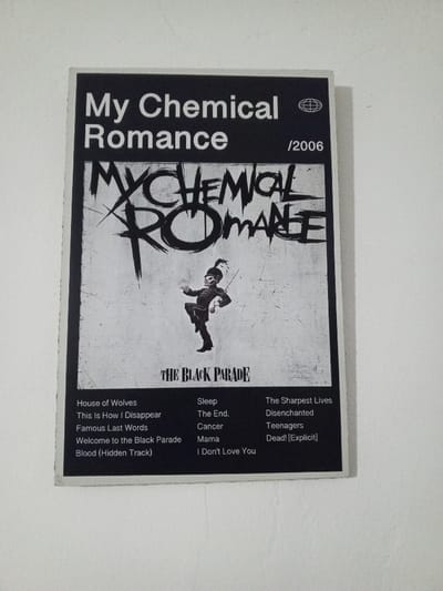 A rectangular, grayscale poster for the musical album "The Black Parade" by My Chemical Romance is affixed to a plain white wall in the city of Al Haouafate, Morocco. The poster's upper section displays "My Chemical Romance" in sans-serif text, a globe icon, and "/2006". Below this, a large, stylized, distressed "MY CHEMICAL ROMANCE" logo dominates the central area. Beneath the logo, a marching skeletal figure, depicted in a band uniform with a shako hat and holding a baton, is centered. The album title "THE BLACK PARADE" is positioned below the figure. The bottom section of the poster lists fifteen song titles in three columns: "House of Wolves", "This Is How I Disappear", "Famous Last Words", "Welcome to the Black Parade", "Blood (Hidden Track)", "Sleep", "The End.", "Cancer", "Mama", "I Don't Love You", "The Sharpest Lives", "Disenchanted", "Teenagers", and "Dead! [Explicit]". The poster exhibits minor warping along its edges but is otherwise intact and clean. The white wall surface appears uniform and unadorned.