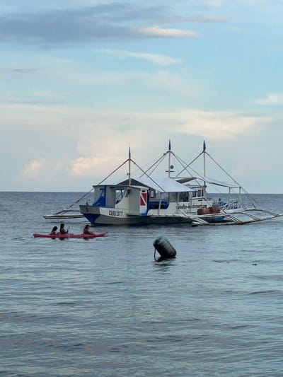 A wide shot captures a tranquil scene on the water near Dauin, Philippines, featuring a boat and a kayak. The sky is a soft gradient of blue and white, with wispy clouds. The sea is a calm, deep blue, with subtle ripples on the surface.
In the mid-ground, a traditional Philippine outrigger boat, known as a "bangka," is moored. The boat is white with blue accents and a red stripe. It has a cabin with windows and a canopy supported by poles. The name "CEBU CITY" is visible on the side of the boat, along with a website address ending in ".com". Attached to the boat are wooden outriggers, providing stability. On the deck of the boat, several cylindrical objects, possibly drums or buoys, are visible.
In the foreground, a bright pink kayak is positioned on the water. Three individuals are seated in the kayak. Two are facing forward, and one is facing backward, appearing to paddle. They are all wearing casual attire.
To the left of the kayak, a dark, cylindrical buoy floats on the water, tethered by a rope. The overall impression is one of a peaceful day at sea, with activities like kayaking and boating taking place.