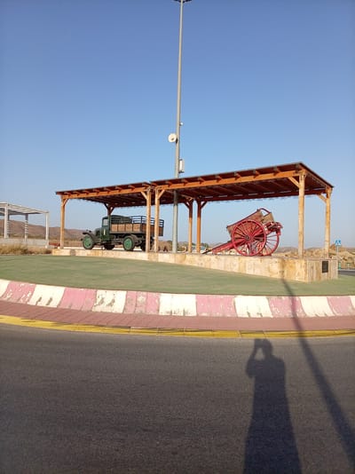 This media file captures an outdoor scene under a clear, bright blue sky, suggesting a sunny day. The primary subjects are two historical vehicles displayed under a wooden pavilion. On the left, a vintage green truck with a wooden bed is parked. To its right, a bright red, two-wheeled cart with large spoked wheels is positioned. Both vehicles are mounted on a raised, light-colored stone platform.

The pavilion itself is constructed with wooden posts and beams, featuring an ornate, patterned edge along the roofline. In the background, there are hints of a semi-arid landscape with low hills or mountains. A tall light pole with antennas stands to the left of the pavilion, and a pedestrian crossing sign is visible in the distance to the right.

In the foreground, a paved road curves into the shot, bordered by a raised planter or curb. This curb has a decorative pattern of pink and white stripes. The shadow of a person taking the photograph is cast onto the road in the lower right portion of the image.

The scene appears to be a public display or monument, possibly in Albox, Spain, given the location context. The overall impression is one of preservation and historical appreciation. The time of day is likely late morning or early afternoon, given the long shadows. No explicit text is visible in the image, apart from the pedestrian crossing sign in the background. The atmosphere is peaceful and somewhat static, highlighting the static nature of the displayed artifacts.