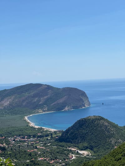A landscape shot focuses on the scenery in Grabovica, Montenegro. The left side of the frame is dominated by a bright, clear blue sky. To the right of the sky, two land masses jut out into the bright blue sea. The land mass on the left side of the frame is a light brown, rugged land mass; it is covered with trees at its base near the water. A boat is present on the water to the right of this land mass. The other land mass on the right of the frame is greener and populated with houses and trees. A beach extends along the coast between the land masses.