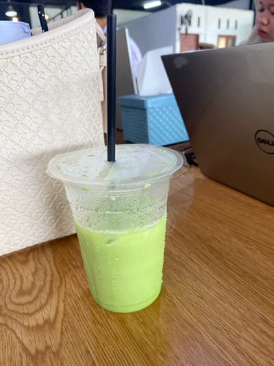 A clear plastic cup, partially filled with a light green, iced beverage and exhibiting condensation, stands on a light brown wooden table. A black straw is inserted through the plastic lid of the cup. To the left, a portion of an off-white, textured handbag with a patterned surface is visible. To the right, an open, bronze-colored Dell laptop is present on the table. Behind the laptop, a light blue, patterned tissue box is situated. A person's head and shoulder, wearing a light-colored headscarf, are partially visible in the upper right background, looking downwards. The scene takes place indoors, likely within a cafe or workspace, in Aluebutuha, Indonesia.