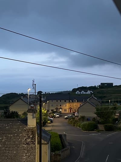 The image depicts a street scene at dusk in Mountcharles, Ireland. In the foreground, the dark, tiled roof of a building is visible, with patches of discoloration or debris. A tall, dark pole stands vertically on the right side of the frame. Beyond this, a street curves to the right. Several houses with dark roofs and yellow-toned walls line the street. Some houses have lit windows, indicating occupancy. A few cars are parked along the street.
Further in the background, a row of houses with uniformly dark roofs and lighter colored walls are situated on a gentle slope. Across the street from these, another cluster of buildings, some with white facades and dark roofs, are visible on a hill. One of these buildings on the hill appears to be a larger, dark-colored structure. The sky is overcast with a gradient of gray and blue tones. Three dark, thin, parallel lines, likely power or telephone cables, stretch horizontally across the upper portion of the image. A communication tower or antenna structure is visible in the mid-ground, to the left of the central street. The overall impression is a quiet, residential area during twilight hours.