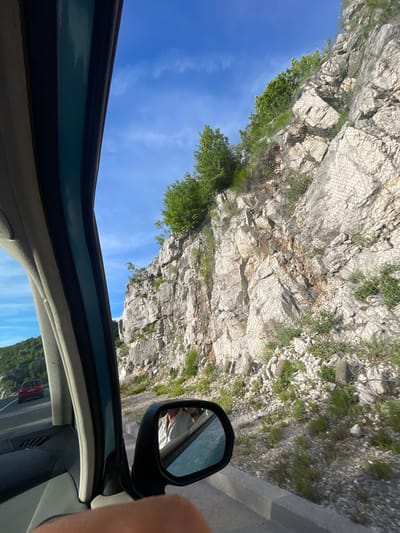 The image captures a view from inside a vehicle traveling on a road adjacent to a rocky mountainside. The perspective is from the passenger seat, looking out the side window. The road appears to be a highway or main route, with another red car visible in the distance traveling in the same direction. The mountainside is characterized by large, light-colored rocks and sparse vegetation, including some trees and shrubs growing from crevices. A metal mesh netting is installed on the mountainside, likely for rockfall protection. The sky is blue with scattered white clouds. A side-view mirror is visible, reflecting a partial image of what appears to be a person inside the car. The interior of the vehicle, including the car's A-pillar and part of the dashboard, is also visible in the foreground, framing the exterior view. The scene appears to be set in a natural, mountainous environment.