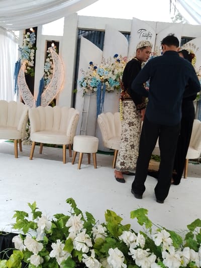This image captures an indoor event setup, likely a wedding ceremony, in Juwana, Indonesia. The scene is dominated by a white aesthetic, featuring a white draped ceiling and a uniform white floor covering.
At the rear, a decorative white backdrop consists of multiple tall, wavy panels, some incorporating dark vertical sections. These panels are adorned with arrangements of white and light blue flowers, interspersed with green foliage. On the left, two large, crescent-shaped white lace decorations, internally illuminated, are mounted vertically. To the right, another floral arrangement, predominantly blue and white, features hanging blue fabric strips. Above the right-hand floral cluster, a white panel displays the calligraphic name "Taufiq."
In the mid-ground, three scallop-backed, cream-colored armchairs with light wooden legs are arranged, along with one matching circular stool.
Three individuals are standing in the center-right of the frame. The central figure, a male, is dressed in traditional Indonesian wedding attire, including a black long-sleeved top with gold embroidery, a patterned brown and cream sarong (batik), and a white, intricately designed headpiece. He stands facing slightly away from the camera. To his right, partially obscuring a female figure, stands a second male, seen from behind, wearing a dark blue long-sleeved collared shirt and black trousers. The female figure, mostly obscured, is wearing a dark garment with visible gold embroidery and dark shoes. The individuals appear to be conversing or standing in preparation for an event.
In the immediate foreground, a low arrangement of white flowers and green leaves is visible.