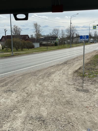 The image captures a daytime scene from inside a moving vehicle, looking forward along a two-lane asphalt road in Bol'shoe Teshkovo, Russia. The road features clear double white lines down its center.

To the left of the road, a green grassy verge separates the roadway from a semi-rural landscape. This area contains several bare trees, suggesting late winter or early spring. Various residential buildings with red and grey roofs are visible beyond the trees. Multiple utility poles with streetlights and overhead power lines line this side of the road. Prominent road signs include two yellow diamond-shaped signs, each depicting a black pedestrian crossing symbol. A blue rectangular sign clearly displays "70" in large white digits, along with the text "РОСАВТОДОР" (ROSAVTODOR), which is the Russian Road Agency, likely indicating a speed limit of 70 km/h or a road designation.

To the right of the road, a wide, unpaved shoulder of dirt and gravel stretches into the foreground, showing textured ground with small stones.

The sky is partly cloudy with bright light, casting faint shadows. A dark interior element with an oval cutout is visible in the bottom left of the frame, confirming the perspective from within a vehicle. The scene depicts a typical, calm road environment.