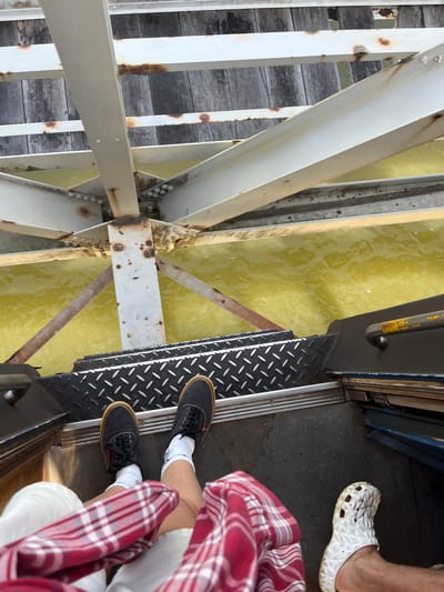 The image shows a low-angle perspective of a person standing on a metal staircase, overlooking murky yellow water. The staircase is constructed from diamond-plate metal, suggesting it is part of a walkway or a vessel. Above the water, a white metal structure with visible rust and bolts spans across, partially obscuring wooden planks. The yellow water below is agitated, indicating movement. The person is wearing dark sneakers with white socks and is partially covered by a red and white plaid garment. Another person's foot, wearing a white slip-on shoe, is visible on the right side of the frame. The scene appears to be outdoors, with elements of a bridge or pier structure visible. The location is identified as Kalutara, Sri Lanka, though specific landmarks are not identifiable from the image alone.