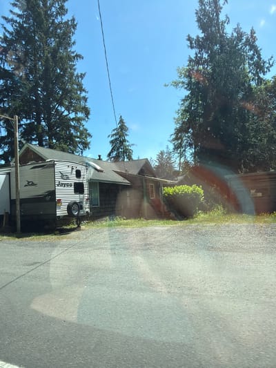 The image depicts a residential scene in Seaside, Oregon, United States, under a clear blue sky. In the foreground, a grey gravel driveway or road is visible. To the left, a white and grey travel trailer, identified by the "Jayco" logo, is parked next to a rustic wooden house with a grey roof. The travel trailer has a spare tire mounted on its rear. The house has a dark wooden exterior and a visible window. Behind the house, tall evergreen trees dominate the landscape, casting shadows on the scene. To the right of the house, another wooden structure with a darker roof and windows is partially visible. Green bushes and some yellow wildflowers grow in front of the structures. A utility pole with wires is also present on the left side of the image. The overall impression is of a somewhat aged, lived-in property surrounded by natural vegetation.