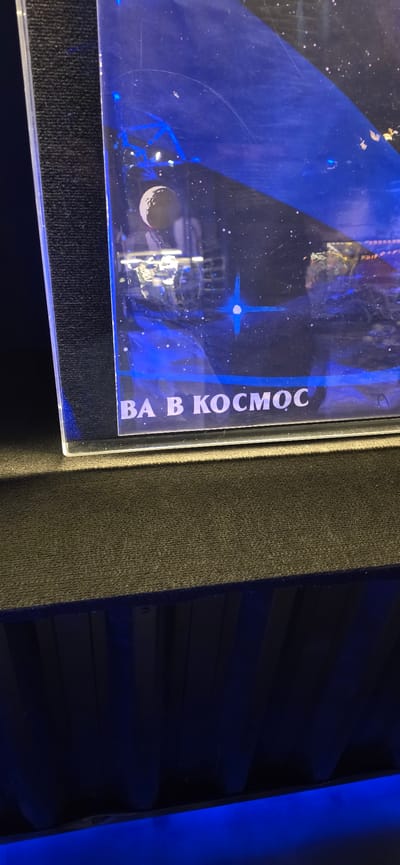 A close-up photograph shows a framed exhibit with artwork depicting space. The artwork features blue tones, celestial bodies, and possibly a rocket or spacecraft design. The Russian text "ВА В КОСМОС" (meaning "Into Space") runs vertically along the right edge of the frame. The exhibit is displayed next to what appears to be a dark gray or black textured surface, possibly part of a larger display case or wall. To the right, a shelf or layered structure is visible, illuminated by a blue light source. The city of Canela, Brazil is not identifiable as the location of this image.